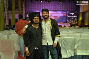 Graduates posing in caps and gowns at ceremony venue with stage in background.