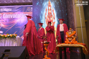 Graduates in red gowns on stage with deity image, statue, and marigold-adorned lamp.