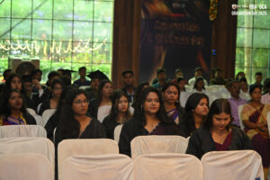 Students in graduation gowns seated at an indoor ceremony with string lights and banner.