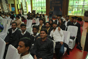 Students in graduation gowns seated indoors, white chairs, windows, banners in background.