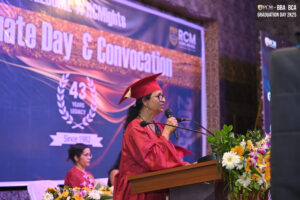 Woman in red cap and gown speaks at RCM graduation event with floral podium arrangement.