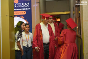 Graduates in red robes and caps with staff in hallway near June 2024 event banner.
