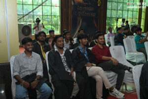 Young men in casual and graduation attire at an indoor graduation event with a large window.