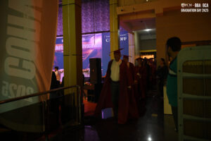 People in maroon graduation gowns and caps walk indoors at a dimly lit event.