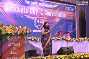 Woman with microphone onstage at Regional College of Management graduation, floral podium, seated officials.