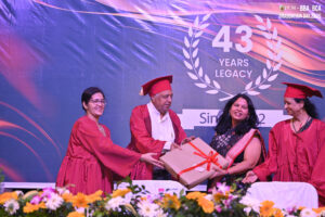 Graduation ceremony with red gowns, woman receives a gift, "43 Years Legacy" backdrop.