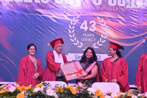 Group in red academic robes at graduation in front "43 Years Legacy" backdrop, exchanging gift.