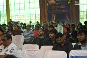Audience at indoor graduation event, wearing casual and formal attire, greenery outside window.