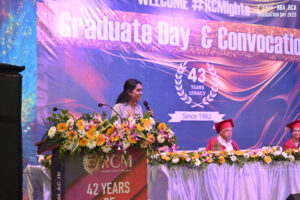 Woman speaks at a podium with flowers at a graduation ceremony; backdrop shows "Graduate Day."