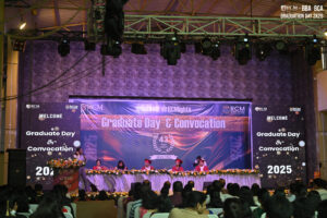 Graduation ceremony with banners, stage, audience, academic robes, and floral decorations.