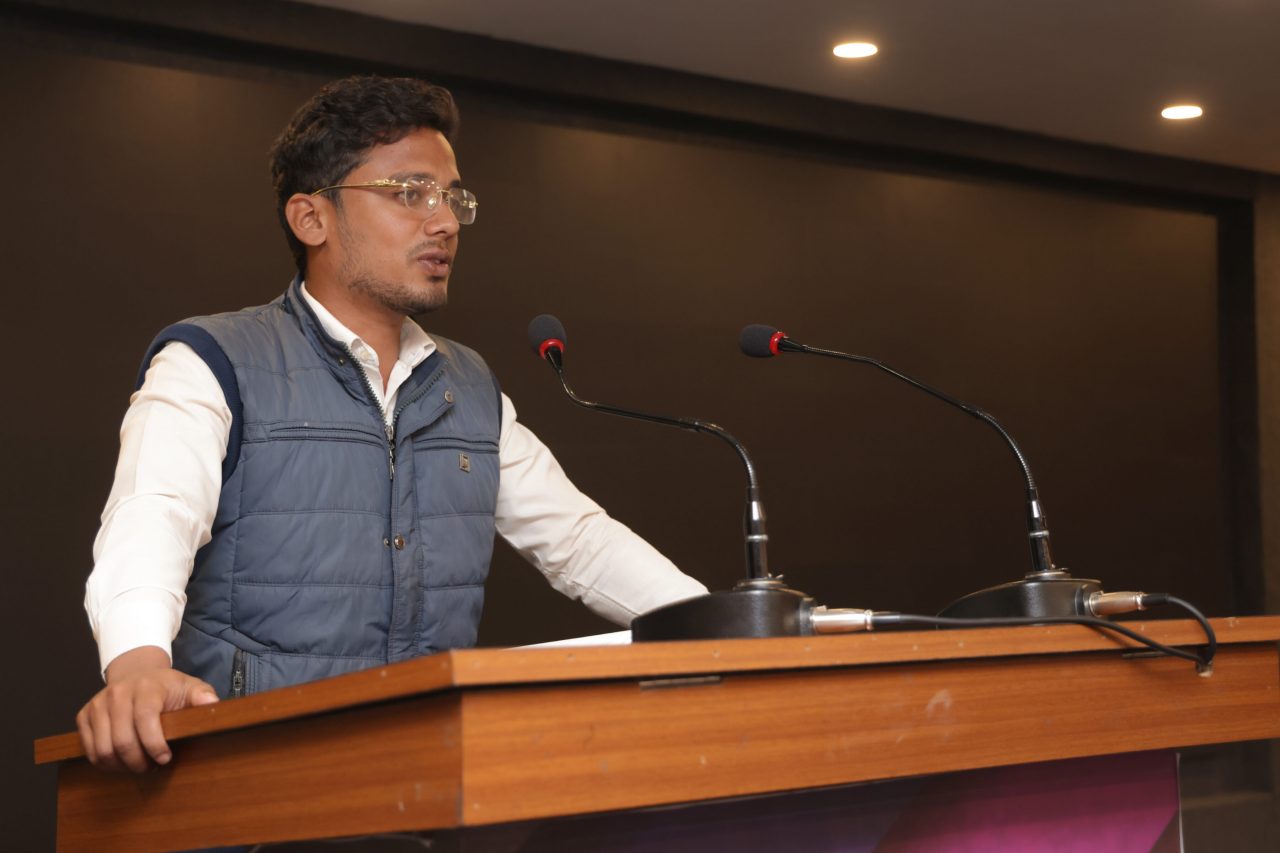 A man in a vest stands confidently at a podium, addressing an audience with a focused expression.